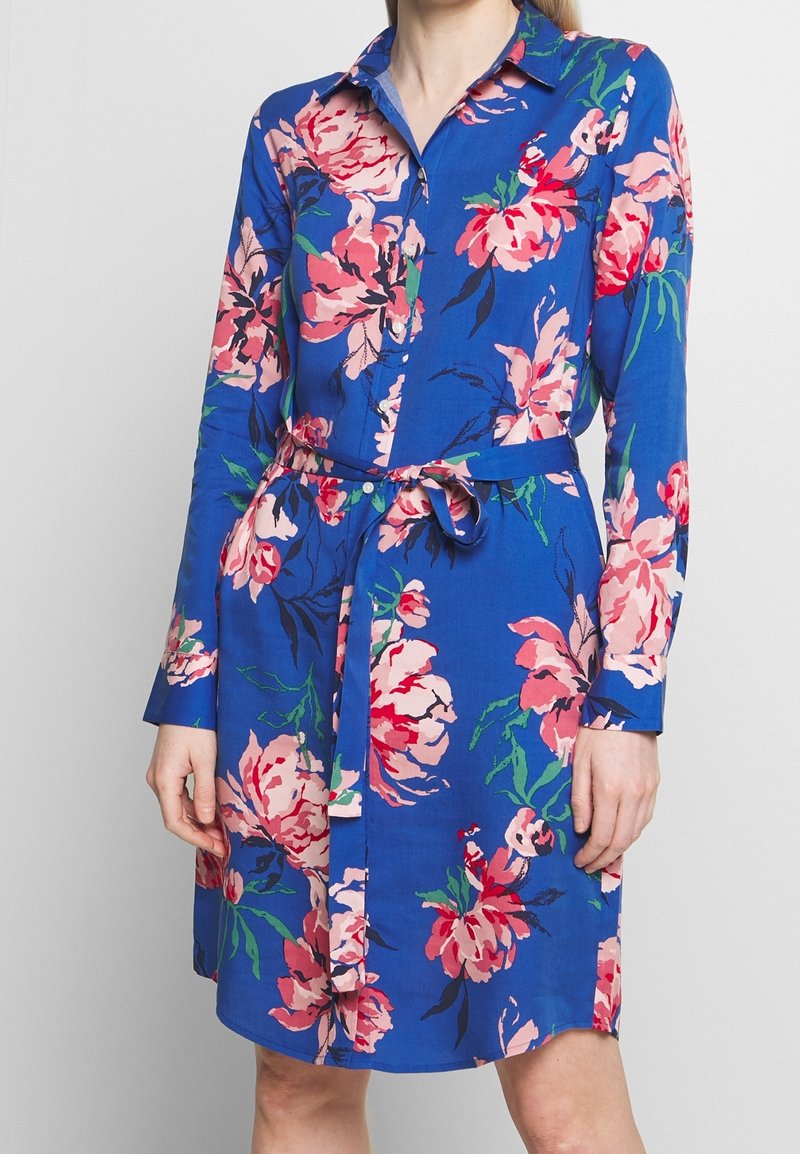 Femme portant une robe bleue à fleurs roses et rouges, manches longues, col, boutonnage devant et ceinture nouée à la taille.