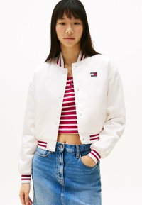 Beskarvet hvid bomberjakke med røde og hvide stribede ribkanter, parret med en rød stribet crop top og denimskørt. Logo på jakken.