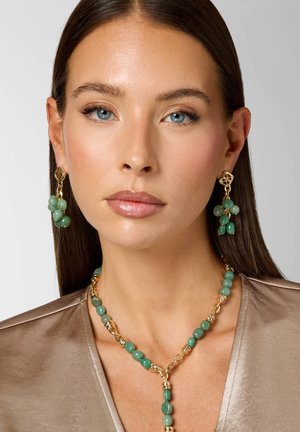 Donna con capelli castani lisci indossa orecchini con perline verdi e catena in oro, e una collana coordinata sopra una camicetta di raso color taupe, su sfondo neutro.