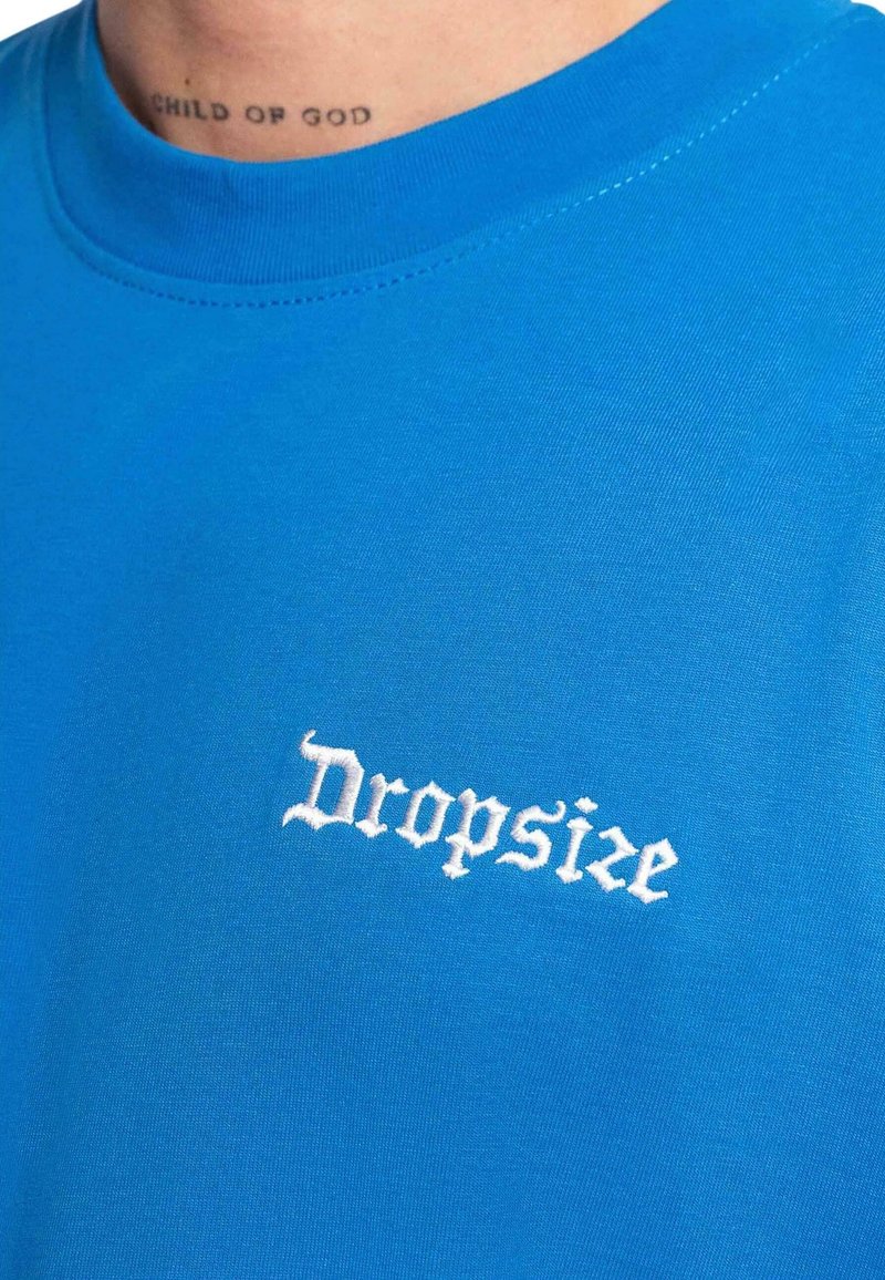 T-shirt bleu avec texte gothique blanc "Dropsize" sur la poitrine et un petit tatouage dans le cou indiquant "CHILD OF GOD".