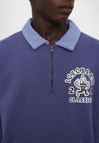 Fialová mikina s svetlomodrým golierom, polovičný zips vpredu a hravá grafika zmrzliny. Text znie "ICECREAM CLASSICS 24." Mäkká textúra.
