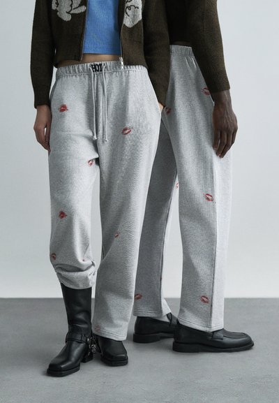 Grijze joggingbroek met rode lippenstiftkuspatronen, voorzien van een taille met trekkoord en taps toelopende boorden, gecombineerd met zwarte enkellaarsjes.