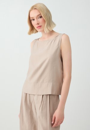 Femme aux cheveux blonds courts portant un haut beige sans manches et un pantalon ample assorti, debout devant un fond clair uni.