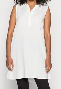 Tunique blanche sans manches avec un col en V et un seul bouton au niveau du col, présentant une coupe ample et un tissu léger et lisse.