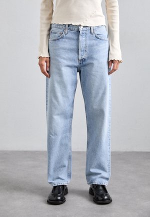 Personne portant un jean droit bleu clair, des chaussures noires et un haut à manches longues crème, debout sur un sol gris devant un mur uni.