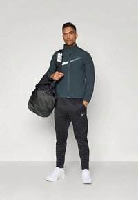 Mørkegrønn zip-up jakke med hvit Nike-logo, sorte sweatpants og hvite sneakers. Bærer en stor svart duffelbag.