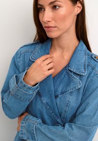 Kvinna med långt brunt hår som bär en blå jeansjacka, rör vid kragen med ena handen och tittar åt sidan mot en enfärgad bakgrund.