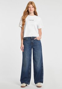 Camiseta corta blanca con el logo "GUESS JEANS", combinada con unos vaqueros de pierna ancha azules. Denim texturizado, costuras sutiles y un ajuste relajado.