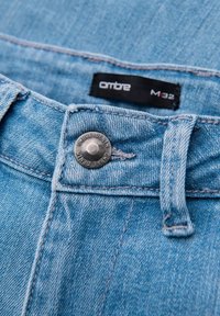 Detail světle modrých džínů z denimu ukazuje kovový knoflík s textem "OMBRE DENIM", poutko na opasek a štítek s velikostí "M 32" na pase.