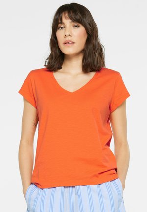 Woman with shoulder-length brown hair wearing an orange V-neck short-sleeve shirt and blue striped pants, standing with hands in pockets.