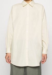 Chemise à manches longues couleur crème avec col pointu, boutonnée sur le devant et coupe ample. Tissu lisse avec un léger éclat.