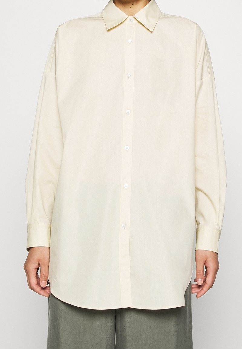 Chemise à manches longues couleur crème avec col pointu, boutonnée sur le devant et coupe ample. Tissu lisse avec un léger éclat.