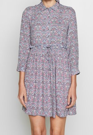 Mujer con un vestido de manga larga hasta la rodilla, con un patrón multicolor de flores y formas geométricas, y una cintura con cordón ajustable.