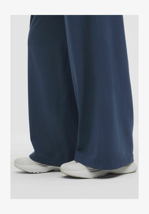 Benen in losse marineblauwe broek en witte sneakers, staande op een witte ondergrond tegen een neutrale achtergrond.