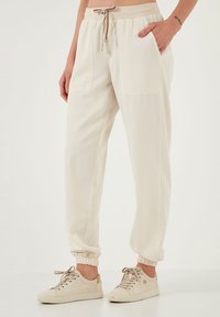 Beige lichtgewicht joggers met elastische tailleband, trekkoord en zijzakken. De boorden zijn elastisch. Draag ze met beige sneakers.