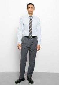 Hellblaues Hemd mit Knopfkragen, graue Anzughose, schwarzer Gürtel und schwarze Schuhe. Die Krawatte hat graue und gelbe diagonale Streifen.