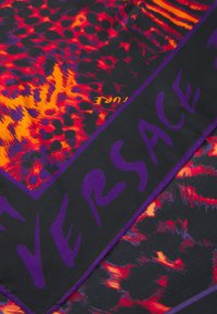 Zīda šalle ar košu rakstu sarkano, oranžo un melno toņu kombinācijā. Malas ir apstrādātas ar violetu, un uz tās ir izcelts uzraksts "VERSACE".