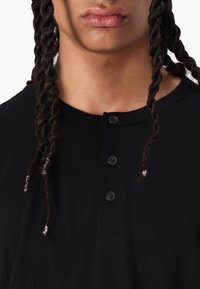 Black cotton t-shirt featuring a round neckline, four button placket, and textured fabric. Braided hair visible on the sides.