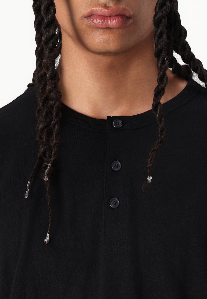 Schwarzes Baumwoll-T-Shirt mit rundem Halsausschnitt, vier Knöpfen und strukturierter Stoffqualität. Geflochtenes Haar an den Seiten sichtbar.