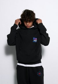 Czarna bluza z kapturem z przednią kieszenią, z wielokolorowym logo "LOCAL HEROES". Gładki materiał, oversize'owy krój, noszona z pasującymi spodniami dresowymi.