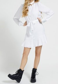 Weißes langärmliges Kleid mit Knopfverschluss, Gürtelbindung in der Taille, ausgestelltem Saum, kombiniert mit schwarzen Kampfstiefeln mit dicken Sohlen.