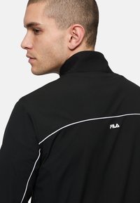 Zwarte jas met een hoge kraag, witte accentstiksel en een klein Fila-logo op de achterkant. Gemaakt van gladde stof met een fitted ontwerp.