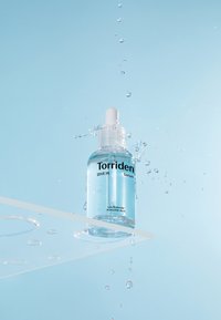 Transparent glasflaske med Torriden serum, udstyret med en hvid pipette, mærket, omgivet af sprøjtende vanddråber mod en blå baggrund.