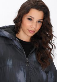 Puffjacke mit einem schwarz-grauen Farbverlauf, einem sternförmigen Reißverschlussanhänger und einem Kapuzenkragen. Glatte, glänzende Textur.