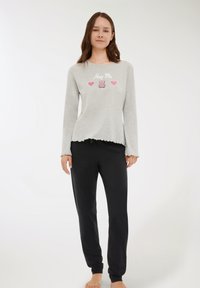 Giovane donna che indossa un top grigio a maniche lunghe con la scritta "Abbracciami" e un orsetto rosa con cuori, abbinato a pantaloni neri larghi, a piedi nudi.