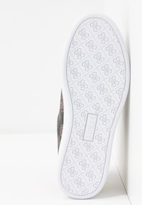Semelle de chaussure blanche avec un motif gaufré répétitif du logo Guess et une étiquette de marque rectangulaire Guess au centre.