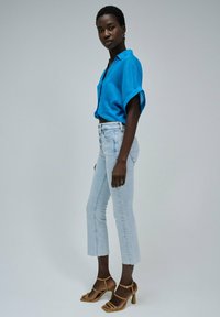 Camisa de lino azul de manga corta, jeans de lavado claro con un corte acampanado, y sandalias de tacón marrones. Fondo liso, enfoque en los detalles de la ropa.