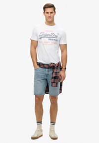 Camiseta blanca de algodón con estampado de "SuperDry", pantalones cortos de mezclilla azul claro, camisa a cuadros roja y azul marino atada a la cintura, y zapatillas crema.