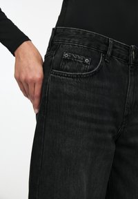 Jean en denim noir à coupe droite, avec une poche avant, des coutures visibles et une ceinture ajustée. Main placée dans la poche.