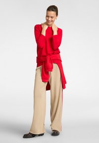 Mujer de pie, vestida con una camiseta de manga larga roja y un sudadera a juego atada a la cintura, pantalones de pierna ancha beige y zapatos marrón oscuro.