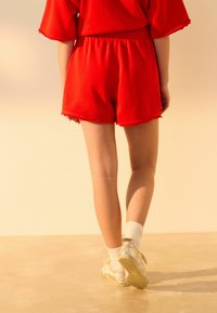 Røde shorts med en afslappet pasform og frynset kant, parret med sølvfarvede sneakers og hvide ankelstrømper. Baggrunden har bløde, varme nuancer.
