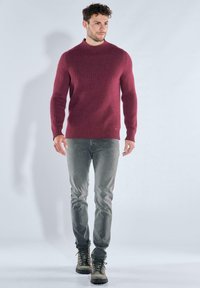 Bordeauxroter, gerippter Pullover mit hohem Kragen, kombiniert mit hellgrauen Jeans und olivgrünen Wanderschuhen. Bodenlanger Schnitt, strukturiertes Gewebe.