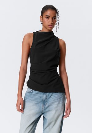 Mujer con cabello rizado, vestida con una camiseta negra sin mangas y vaqueros azul claro, de pie frente a un fondo liso gris claro.