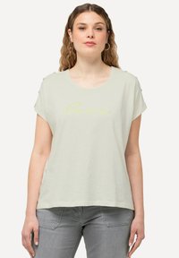 Camiseta de algodón verde claro con escote redondo, mangas cortas y una impresión en cursiva amarilla que dice "cuidado". Presenta botones decorativos en los hombros.