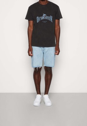 Man wearing black graphic t-shirt, light blue frayed denim shorts, and white sneakers, standing against a plain light wall.