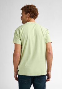 Camiseta de algodón verde claro con cuello redondo y mangas cortas, que presenta una textura sutil. Llevada con jeans oscuros, vista desde la parte trasera.