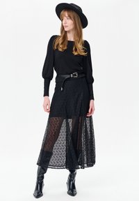 Zwarte top met lange mouwen gecombineerd met een doorschijnende zwarte kanten rok met bloemmotieven, getailleerd met een zwarte riem; model draagt een zwarte hoed.