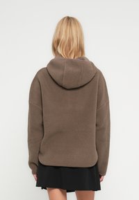 Γυναίκα με ξανθά μαλλιά που φοράει καφέ φλις ζακέτα με κουκούλα και μαύρη φούστα, αποθανατισμένη από πίσω μπροστά σε απλό λευκό φόντο.