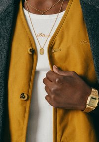 Lähikuva henkilöstä, jolla on päällään sinappinkeltainen takki, valkoinen raidoitettu paita, kaksi kultaketjua ja kultakello, pitäen takin lievettä.