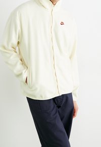 Vit fleecejacka med hög krage, framzipper och röd Nike-logotyp på bröstet; har två sidofickor.