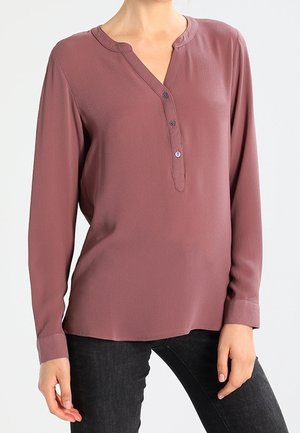 Vrouw draagt een mauve blouse met lange mouwen en een drieknops sluiting, en zwarte broek tegen een effen lichte achtergrond.