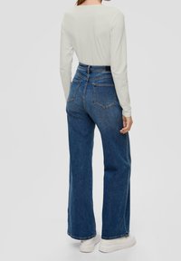 Jeans denim biru dengan pinggang tinggi dan kaki lebar, desain lima kantong, dan sulaman halus. Dipadukan dengan atasan lengan panjang ringan dan sepatu putih.