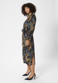 Robe fleurie en tissu noir, présentant des motifs jaunes et bleus, manches longues, fentes latérales et taille ceinturée. Associée à des talons noirs.