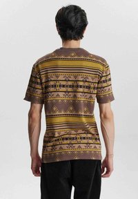 Camiseta de manga corta marrón con patrones geométricos en amarillo y negro. Presenta un cuello redondo y un corte holgado, adecuado para un estilo informal.