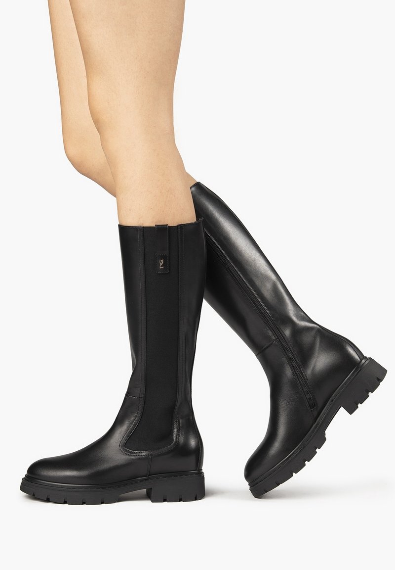 Botas negras hasta la rodilla hechas de cuero suave con paneles laterales elásticos y suela de goma texturizada, con una cremallera lateral para facilitar su uso.