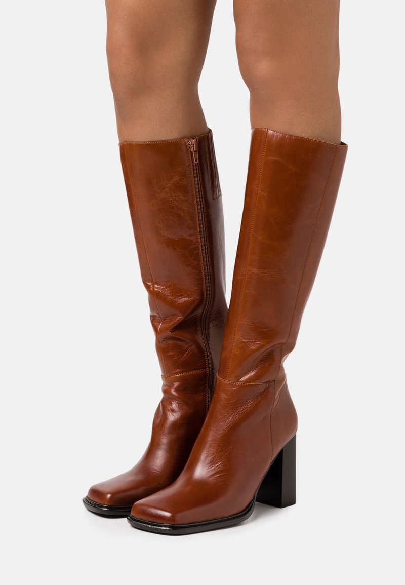 Bottes en cuir marron montantes avec un bout carré, une finition brillante et un talon épais noir. Équipées d'une fermeture éclair sur le côté pour un enfilage facile.